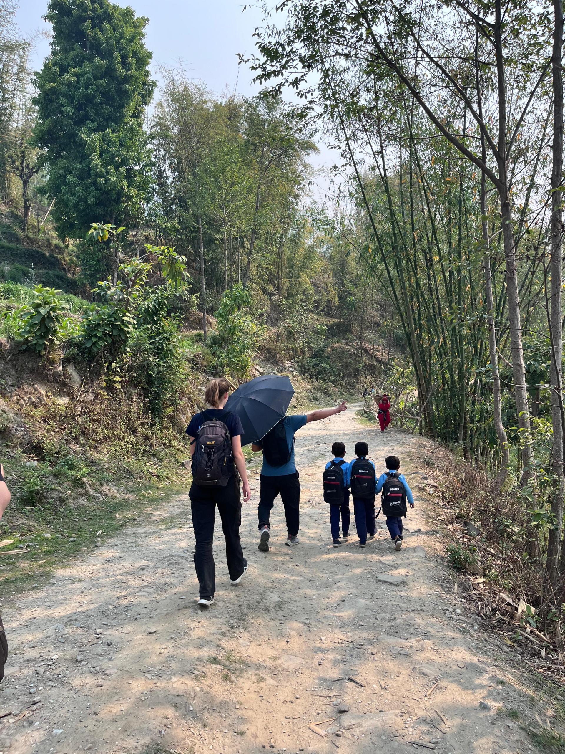 2 Menschen und 3 Kinder (von hinten) auf dem Weg zur Schule. Einer hält einen Regenschirm