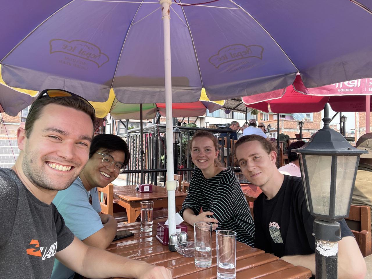 Vier Menschen sitzen unter einem Sonnenschirm an einem Tisch und lachen alle in die Kamera