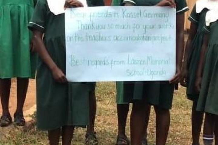 Schulkinder der Lauren Patterson Memorial School halten ein Schild hoch mit der Aufschrift "Dear friends in Kassel, Germany, thank you so much for your work on the teachers accomodation project. Best regards from Lauren Memorial School Uanda"