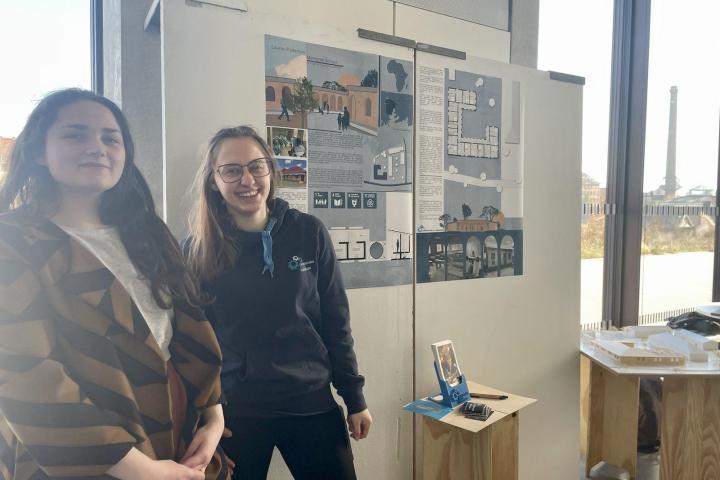 zwei lächelnde Mitglieder des IOG-Teams stehen in der Universität Kassel neben Plakaten mit Informationen und Planzeichungen zur Lauren Patterson Memorial School